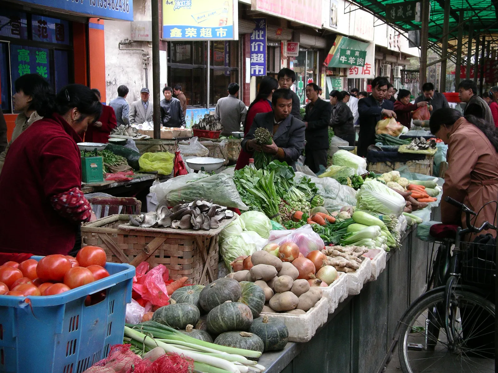 A street produce market in China, vegetable stalls lining a narrow lane with stallholders and shoppers.